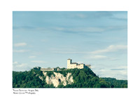 A landscape photograph of Angera Fortress as seen from Arona, featuring a clear blue summer sky and a few white clouds.