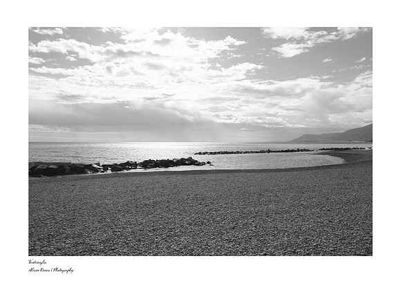 Artistic black-and-white photograph of the beach at Ventimiglia in Liguria, featuring rocks, cliffs, and a contrasting cloudy sky.