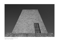 A fine art black-and-white print of the Littoria Tower in Carbonia. A low-angle shot that emphasizes the monumentality of the Rationalist architecture, featuring a dark, high-contrast sky and sharp ge
