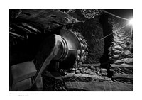 Black-and-white fine art photograph of industrial machinery inside the Carbonia coal mine in Sardinia. A starburst of light illuminates the darkness on the right, creating a stark contrast with the me