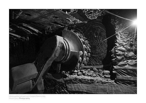 Black-and-white fine art photograph of industrial machinery inside the Carbonia coal mine in Sardinia. A starburst of light illuminates the darkness on the right, creating a stark contrast with the me